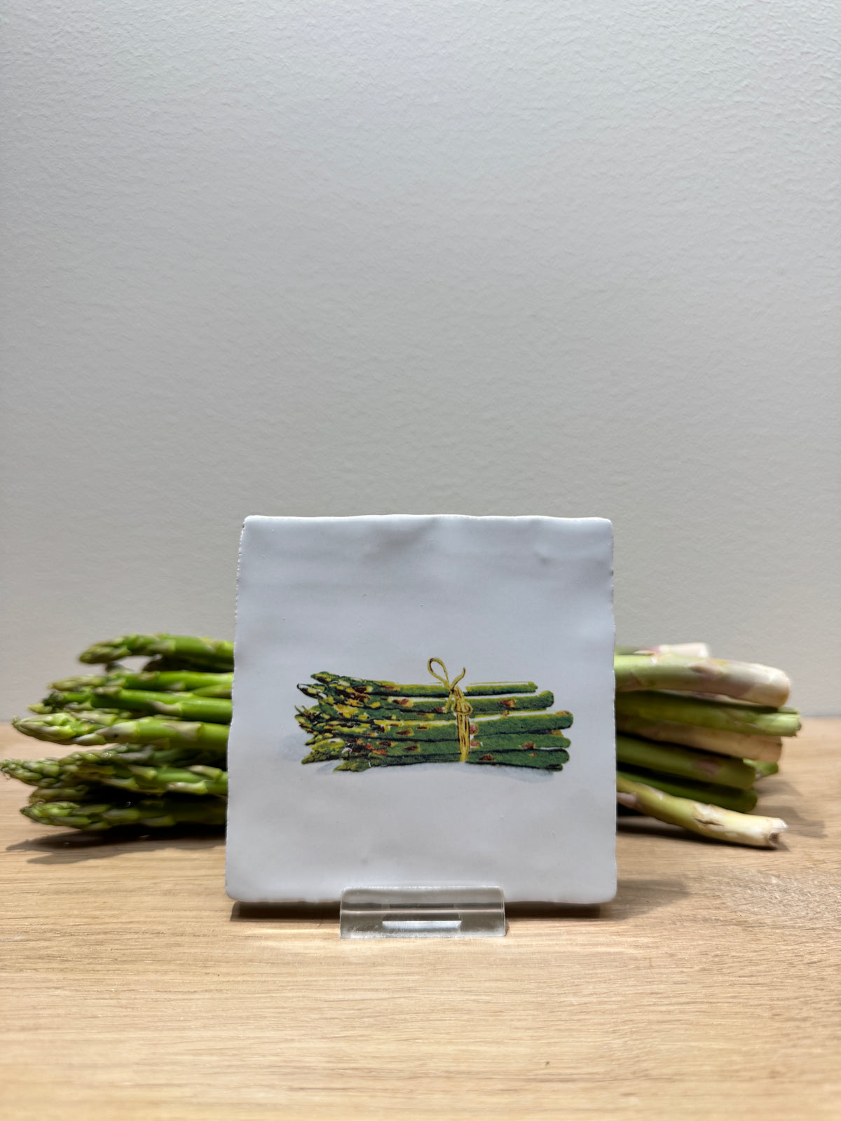 A square ceramic coaster with a hand-painted illustration of a green asparagus bundle tied with a yellow string, displayed on a clear acrylic stand on a wooden table with asparagus props.