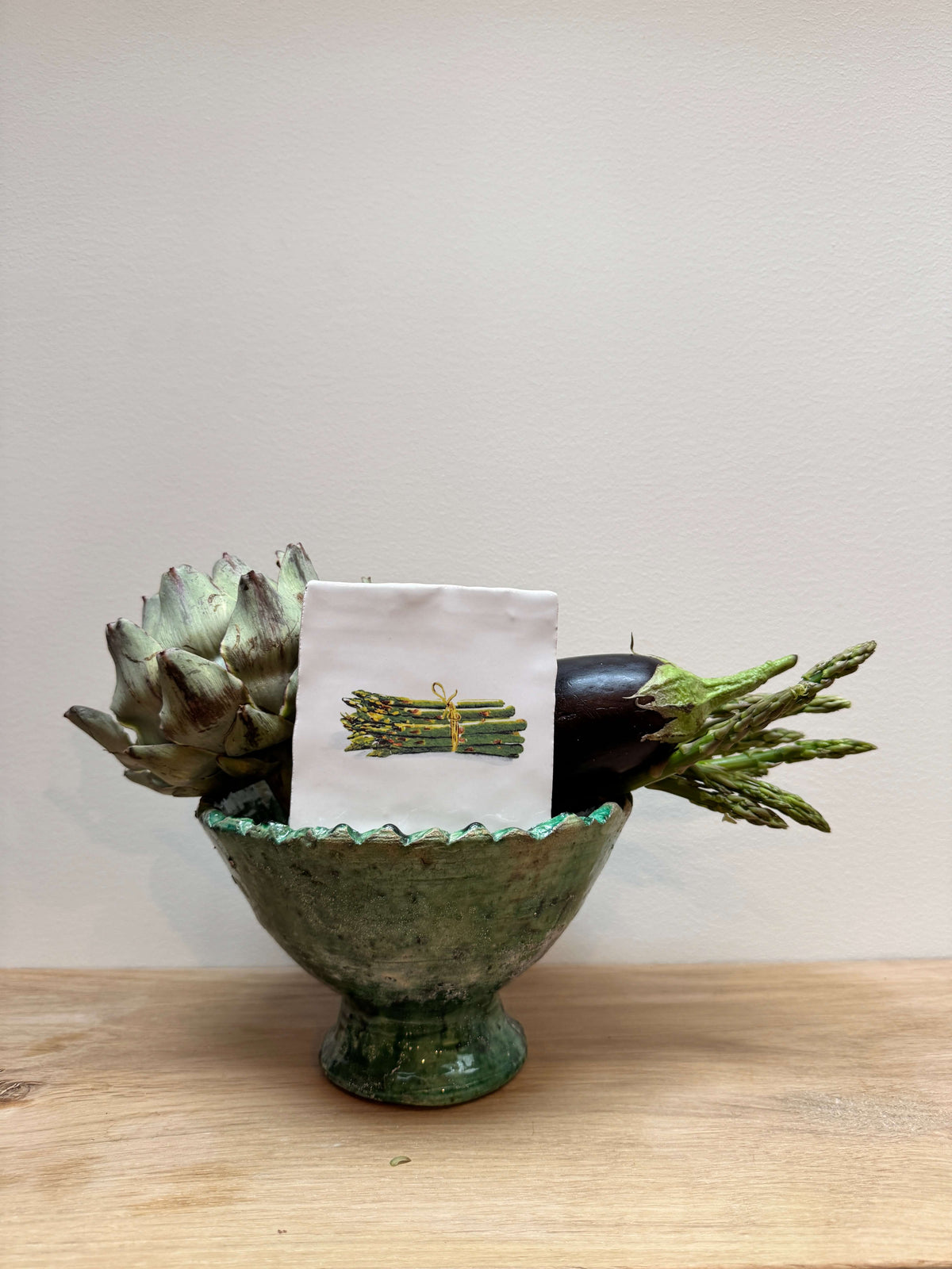 A small green ceramic pedestal bowl filled with decorative vegetables, including an artichoke, eggplant, and asparagus, with a small white card featuring a green and yellow illustration of a bundle of asparagus in the center, set on a light wood surface against a plain beige wall.