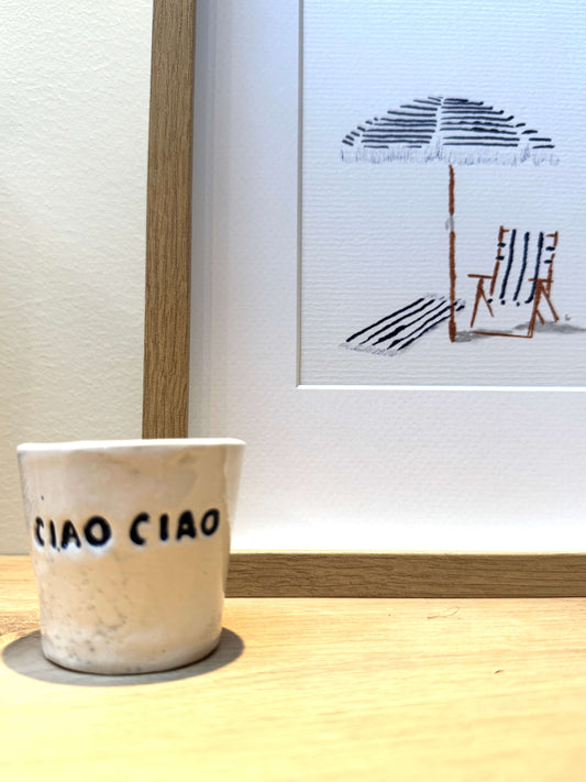 A small speckled beige ceramic cup with the words 'CIAO CIAO' printed in blue on the front, sitting on a light wood surface in front of a framed beach-themed print.