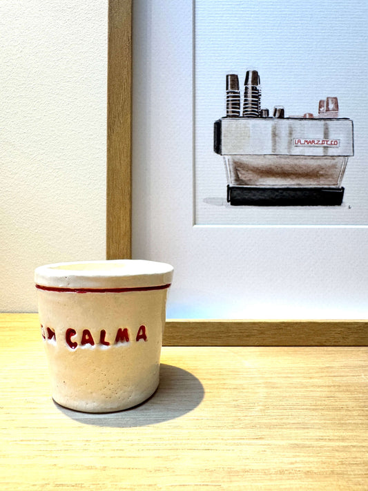 A small beige ceramic cup with red lettering reading 'CON CALMA' sits on a light wood surface in front of a framed print showing a La Marzocco espresso machine.
