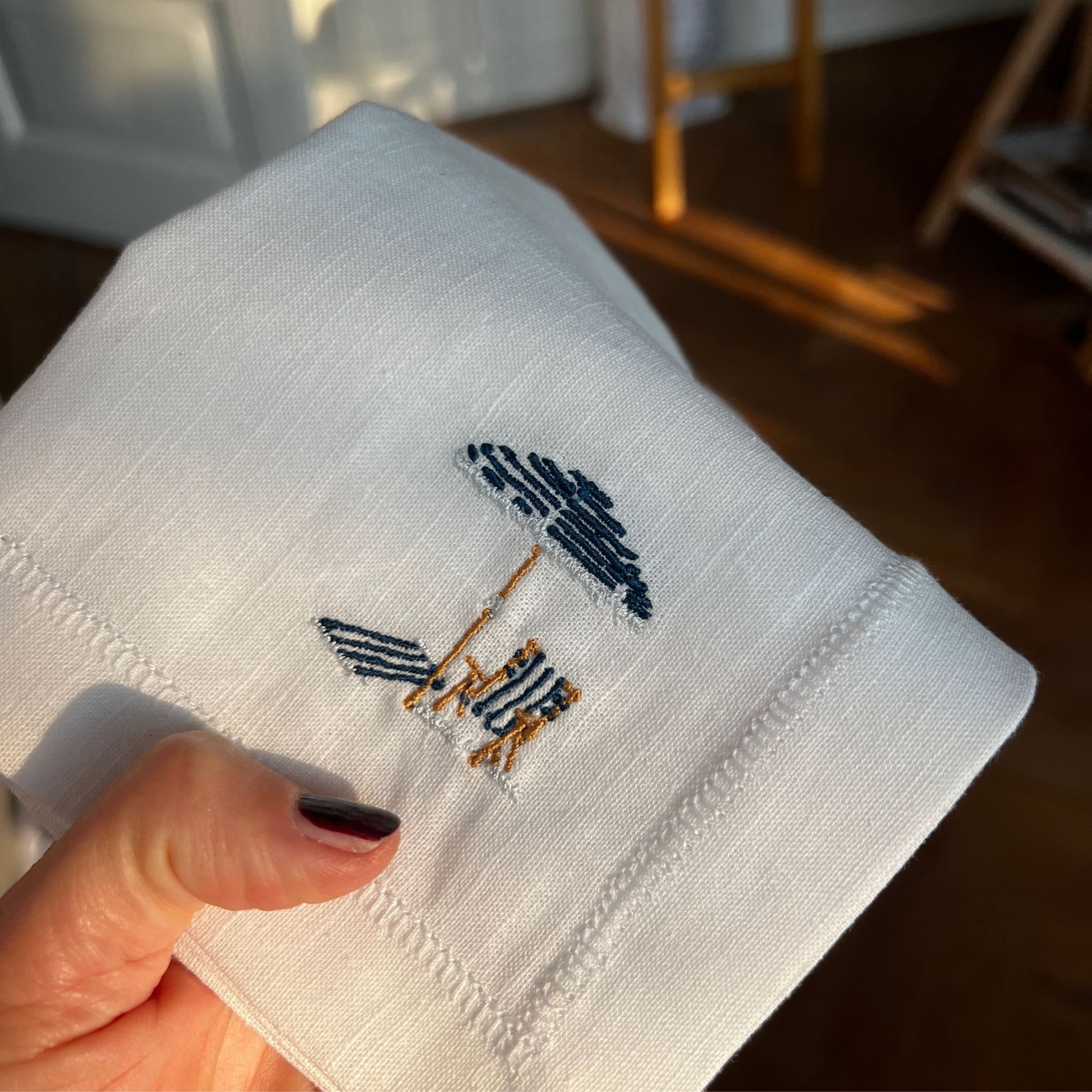 Detailed embroidery of illustration by Stefanie Viëtor of blue parasol on white linen napkin with hemstich of 40x40cm
